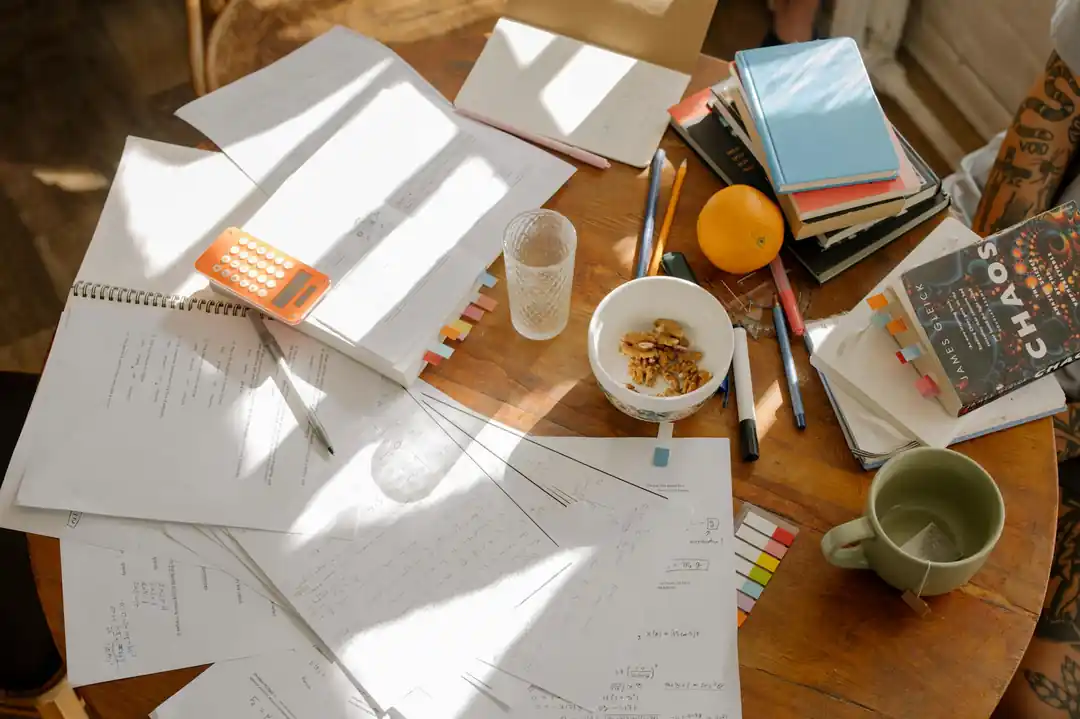 A cluttered study table with books, notes, and study materials in natural light.