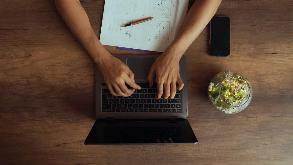 A person typing on a laptop from above, surrounded by papers and a smartphone on a wooden table.