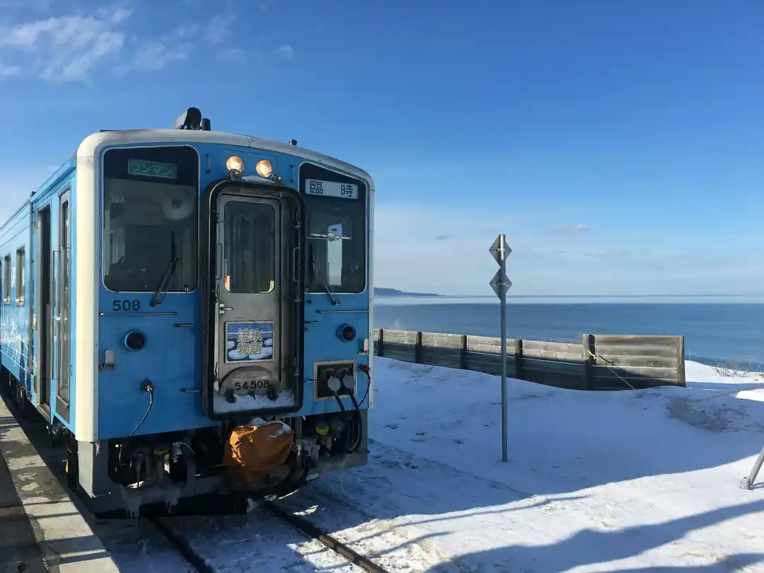 雪景色の海岸線の隣を走る青い電車。冬の厳しい寒さと、列車の暖かさの対比が感じられる。