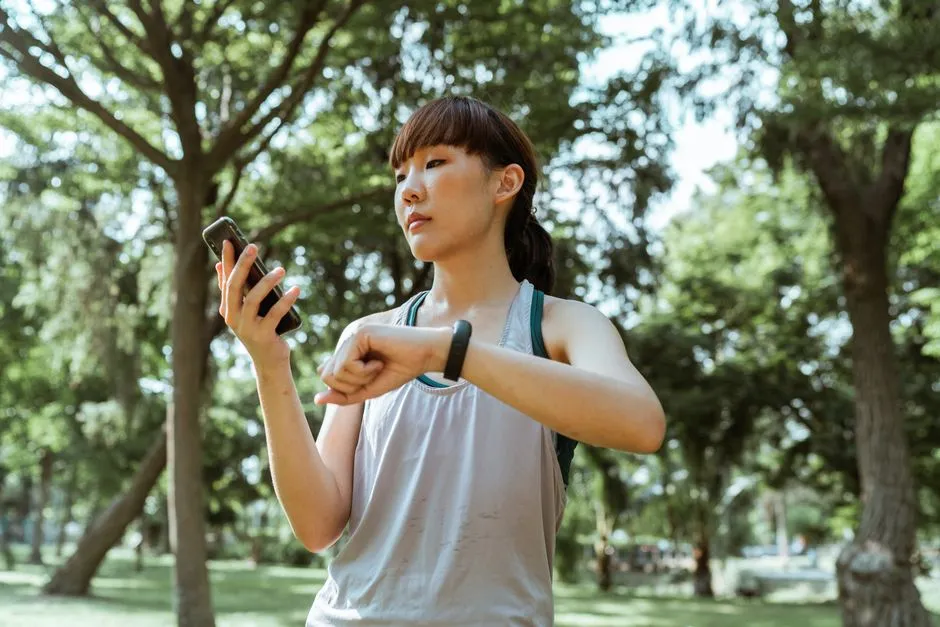 一位身穿運動服的年輕亞洲女性在陽光公園中查看手機與健身手環