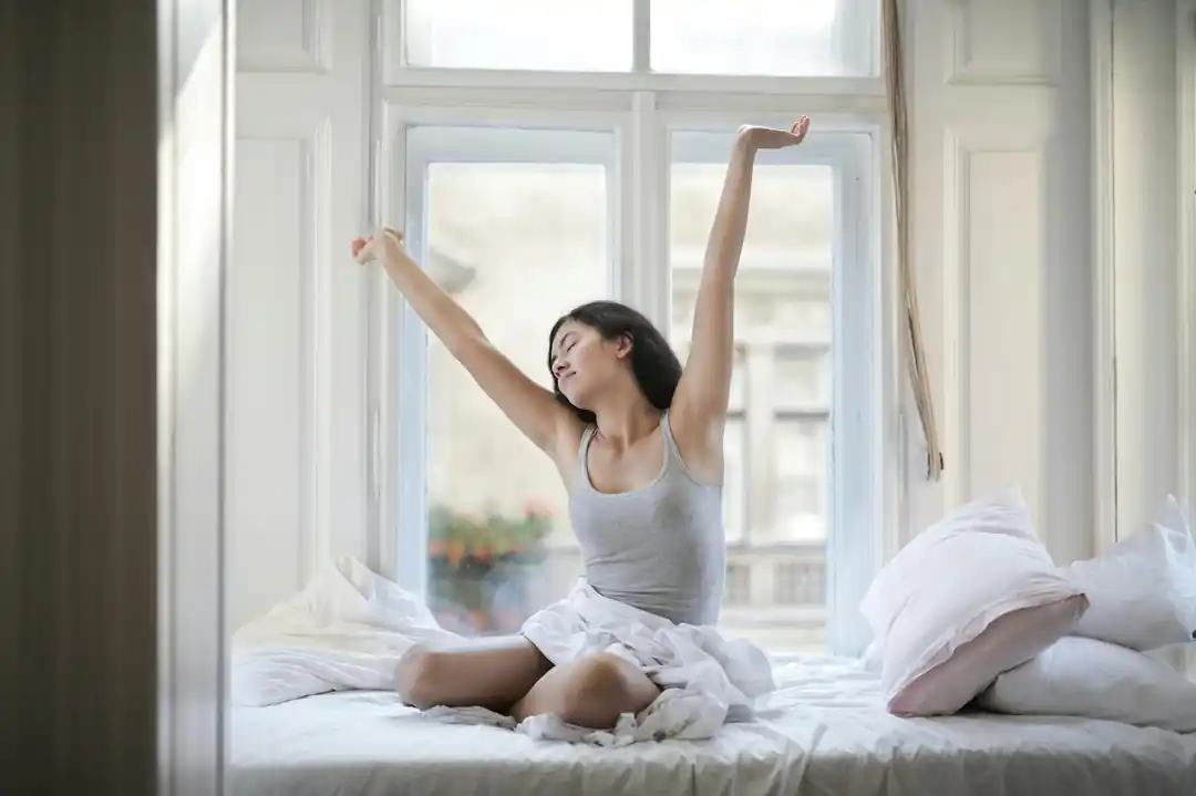 明るい部屋のベッドの上で、気持ちよさそうに伸びをするグレーのタンクトップを着た女性。