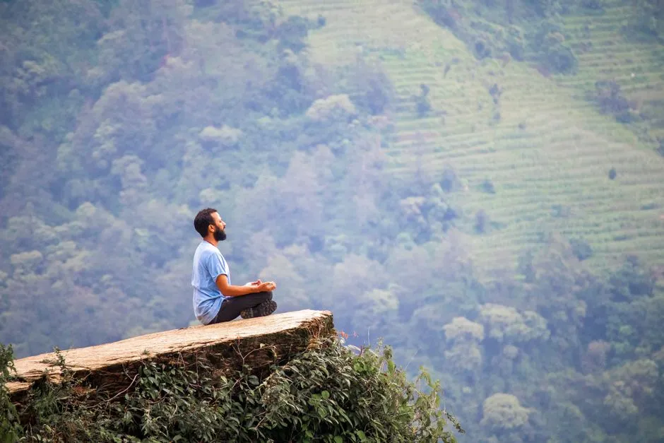 A man meditating on a cliff with lush green mountains in the background.