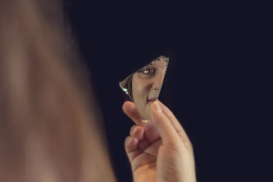 A person's eye and face reflected in a shard of a broken mirror against a dark background.