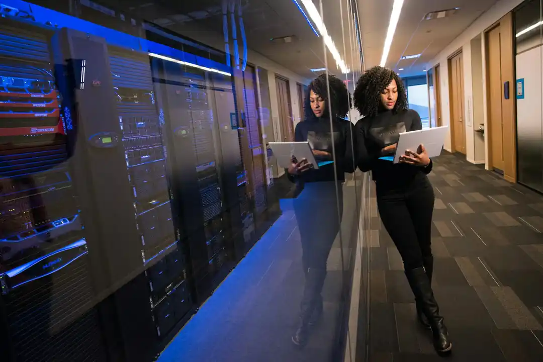 A woman in a data center hallway, working on a laptop in front of server racks.