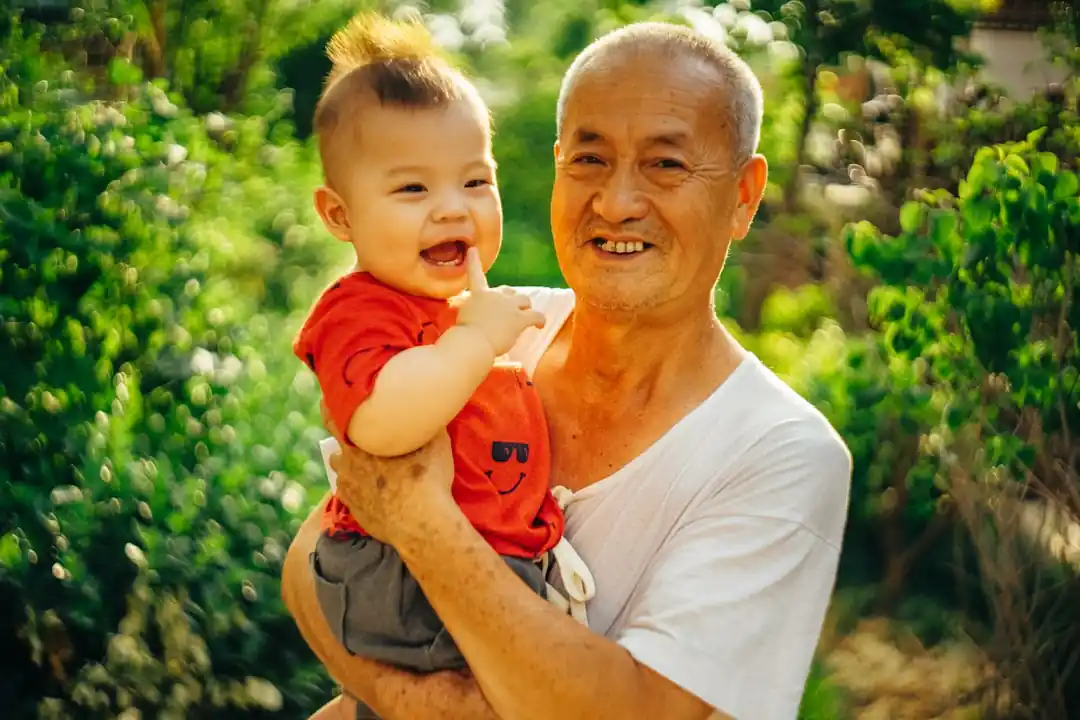 一位年長的亞洲男性抱著一個嬰兒,站在綠色植物前,臉上洋溢著幸福的笑容。