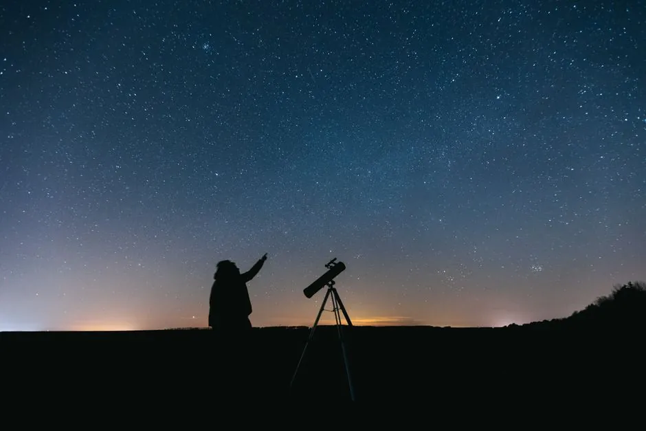 A silhouetted person standing with a telescope under a bright starry night sky